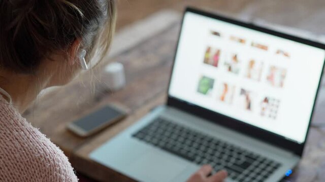 young unrecognizable woman with wireless earphones sits at wooden desk at home, browsing site of online store on laptop and making online order from home by phone. focus on earphone