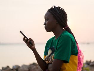 Young beautiful black girl using smart phone on sea at sunset