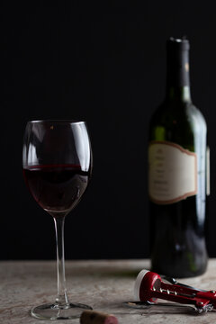Image of a standard size wine glass with red wine filled halfway through. There is a corkscrew on the marble countertop with a cork lying beside. A green glass bottle sits in the dark background.