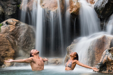 Fototapeta na wymiar Wellness spa couple freedom at waterfall relaxing with open arms carefree. People enjoying water falling in natural pool in tropical nature background. Healthy lifestyle.