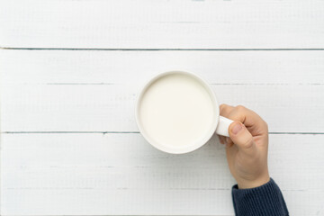 Child hand holding cup of milk against white wood background, top view.