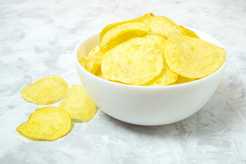 Golden solted natural potato chips in white ceramic bowl on concrete background.