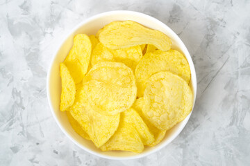 Natural potato chips in white ceramic bowl on textured grey background, top view.