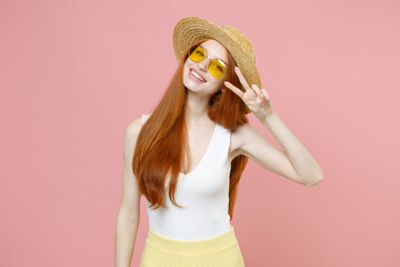 Young smiling happy redhead cute gladden cheerful woman 20s ginger long hair wearing straw hat glasses summer clothes show victory v-sign gesture isolated on pastel pink background studio portrait.