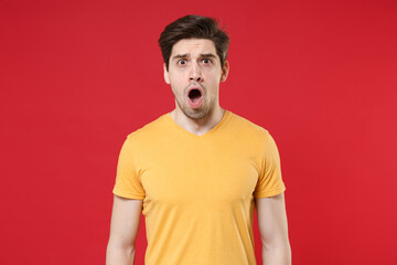 Young shocked confused unshaved caucasian impressed man 20s years old in casual basic blank print design yelow t-shirt look camera with open mouth isolated on red color background studio portrait