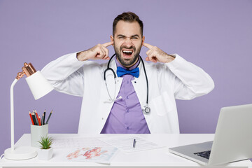 Irriatated nervous male doctor man wearing white medical gown suit sit at desk work on computer in clinic office cover ears with fingers shout scream isolated on violet background studio portrait