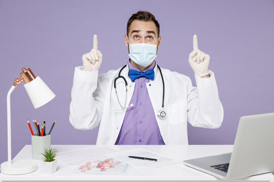 Male Doctor Man In White Medical Suit Face Mask Gloves From Coronavirus Covid-19 Sit At Desk In Clinic Office Point Index Fingers Overhead On Copy Space Isolated On Violet Background Studio Portrait.