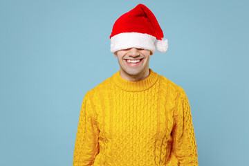 Young funny cheerful smiling man 20s in casual knitted yellow touching sweater put a hat over your eyes santa hat celebrating christmas winter holidays isolated on blue background studio portrait.