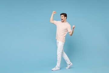 Full length of young caucasian happy excited successful student man 20s in beige t-shirt white pants doing winner gesture clenching fists look aside isolated on blue colorbackground studio portrait.