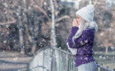 Side view young woman warming her hands, wears warm clothing on snowing winter day outdoors. Cute girl feeling cold. Winter concept.