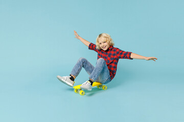 Full length of cheerful little curly kid boy 10s in basic red checkered shirt sitting on skateboard spreading hands isolated on blue background children studio portrait. Childhood lifestyle concept. © ViDi Studio