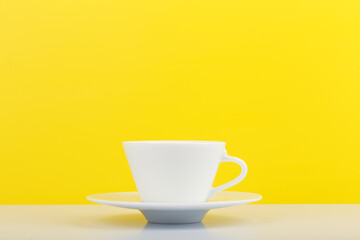 Minimalistic still life with white glossy cup of coffee on white table against yellow background with copy space. 