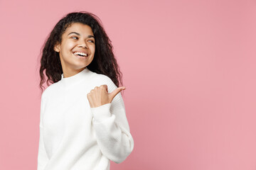 Young smiling happy african american curly woman 20s in white knitted sweater point thumb finger back on copy space workspace mock up look aside isolated on pastel pink background studio portrait.