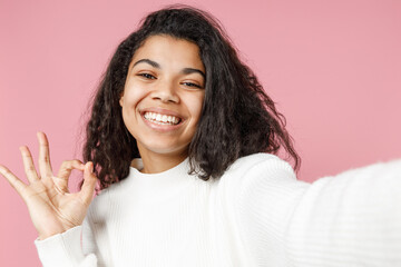 Close up young happy positive cheerful african american curly woman 20s in white knitted sweater do selfie shot on mobile phone show ok okay gesture isolated on pastel pink background studio portrait