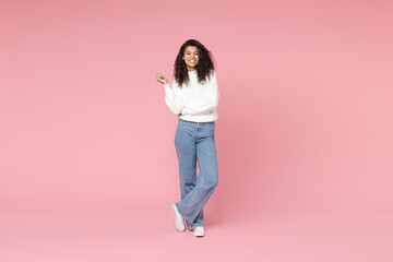 Full length young smiling cute african american woman 20s curly hair wear white casual knitted sweater jeans look camera hold hands crossed folded isolated on pastel pink background studio portrait