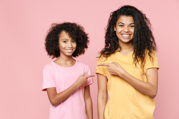 Funny african american young woman and little kid girl sisters wearing basic t-shirts pointing index fingers at each other isolated on pastel pink color background studio portrait. Family day concept.