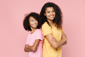 Smiling funny african american young woman little kid girl sisters in casual t-shirts standing back to back looking camera holding hands crossed isolated on pink background studio. Family day concept.