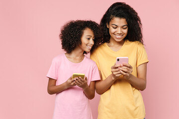 Smiling african american young woman and little kid girl sisters in casual t-shirts using mobile cell phone typing sms message isolated on pastel pink background studio portrait. Family day concept.