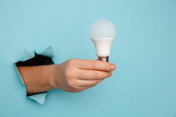 Hand holding an incandescent led light bulb from a torn hole in blue paper.