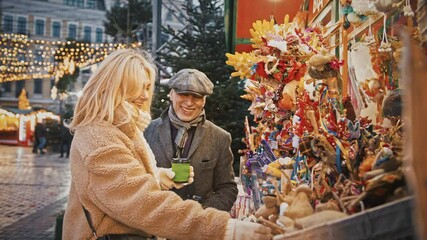 Outdoor portrait of happy senior man and woman enjoying Xmas fair in city center, walking with coffee, slow motion - Powered by Adobe