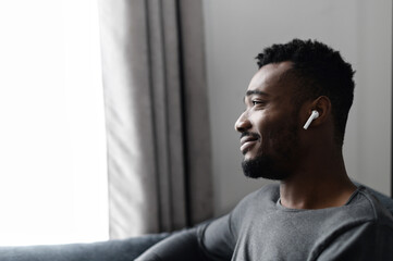 Close-up side view on handsome African-American guy with an airpods earphones spends time at home,...