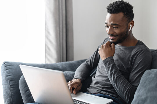 A cheerful smiling African-American guy sits with laptop on comfortable couch and types on the keyboard, answering email, chatting in networks