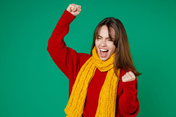 Overjoyed young brunette woman 20s in casual knitted red sweater yellow scarf doing winner gesture clenching fists celebrating keeping eyes closed isolated on green color background studio portrait.