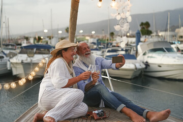 Senior couple toasting champagne while taking selfie on sailboat vacation - Happy mature people having fun celebrating wedding anniversary on boat trip - Love relationship and travel lifestyle concept