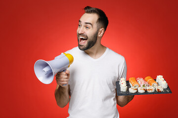 Excited young bearded man 20s wearing basic white t-shirt screaming in megaphone hold makizushi sushi roll served on black plate traditional japanese food isolated on red background studio portrait.