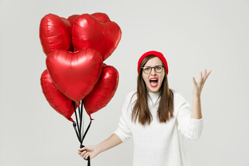 Angry young woman in sweater red hat glasses spreading hands screaming swearing celebrating birthday holiday party hold bunch of heart air inflated helium balloons isolated on white background studio.