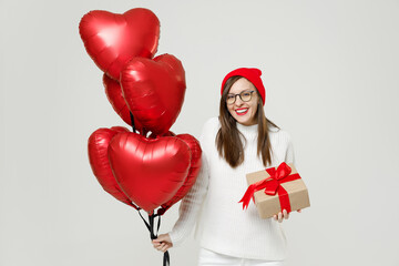 Fototapeta na wymiar Funny young woman in sweater red hat glasses celebrating birthday holiday party hold present box with gift ribbon bow bunch heart air inflated helium balloons isolated on white wall background studio.