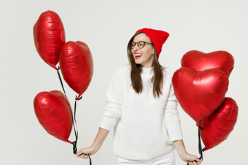 Laughing young brunette woman in sweater red hat glasses looking aside celebrating birthday holiday party hold bunch of heart air inflated helium balloons isolated on white background studio portrait.