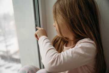 Fototapeta na wymiar Little girl in pink pajamas sits near the window. Quarantine. Lockdown.