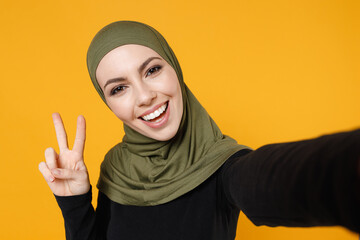 Close up of excited young arabian muslim woman in hijab black green clothes doing selfie shot on mobile phone showing victory sign isolated on yellow background. People religious lifestyle concept.