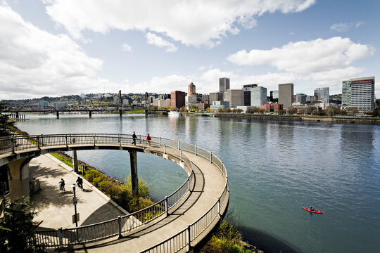 Looking over the Eastbank Esplanade towards downtown Portland on a sunny day.  A handful of people are walking in the foreground and a kayaker rows by in the river.