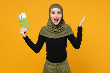 Surprised traveler tourist arabian muslim woman in hijab black green clothes hold passport tickets spreading hands isolated on yellow background studio portrait. People religious lifestyle concept.