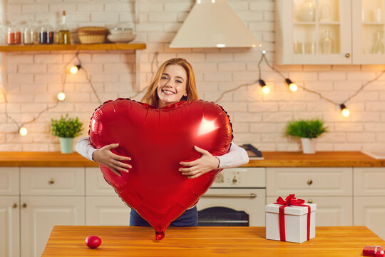 Portrait of a joyful excited young girl hugging a big red heart shaped balloon looking at the camera. Girl stands at home in the kitchen and receives gifts for Valentine's Day from her lover.