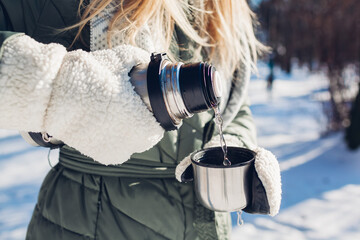 Woman pouring hot tea out of vacuum flask in winter park. Drinks to warm up in snowy frosty weather...