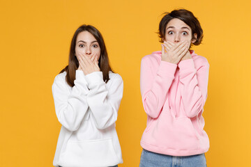 Shocked surprised two young women friends 20s wearing casual basic white pink hoodies standing covering mouth with hands looking camera isolated on bright yellow color background studio portrait.