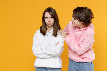 Displeased offended smiling funny two young women friends 20s wearing casual white pink hoodies holding hands crossed hugging apologizing isolated on bright yellow color background studio portrait.