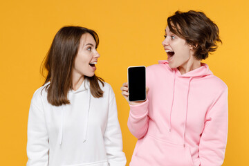 Shocked excited two young women friends 20s wearing casual white pink hoodies hold mobile cell phone with blank empty screen looking at each other isolated on bright yellow background studio portrait.