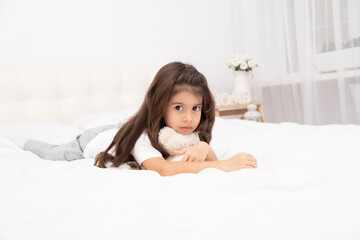 sad little brunette girl lying with teddy bear on bed at home