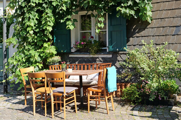 Fototapeta na wymiar Close-up of a cute table with a bench and chairs in front of a slate plated house with green shutters and decorated with plants