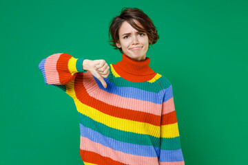 Displeased confused upset young brunette woman 20s years old wearing casual basic colorful sweater stand showing thumb down looking camera isolated on bright green color background studio portrait.