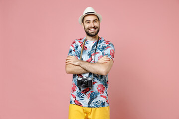 Smiling young traveler tourist man in summer clothes hat with photo camera hold hands crossed isolated on pink background. Passenger traveling abroad on weekends getaway. Air flight journey concept.