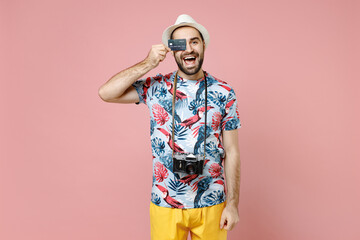 Excited young traveler tourist man in summer clothes hat covering eye with credit bank card isolated on pink color background studio. Passenger traveling abroad on weekend. Air flight journey concept.