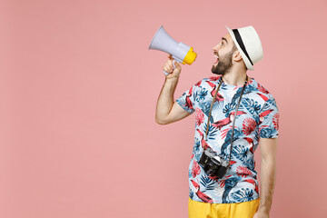 Excited young traveler tourist man in summer clothes hat screaming in megaphone looking aside isolated on pink background studio portrait. Passenger traveling on weekends. Air flight journey concept.