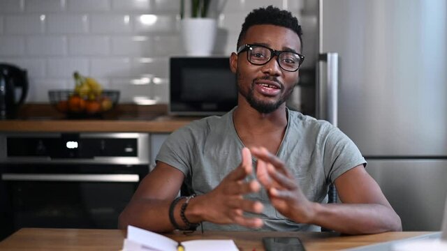 Video screen view of smiling African-American man sits at the desk in the kitchen at home talk on video call with employee, friends. Multiracial male have video conversation, virtual meeting