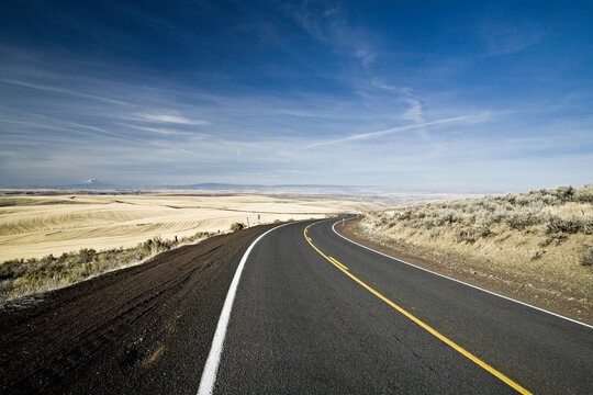 A single lane highway curves around a very low hill in the high desert of Eastern Oregon.  A dry, arid landscape stretches on for miles in the distance.
