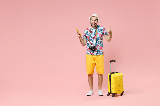 Full length of excited young traveler tourist man using mobile phone booking hotel taxi showing thumb up isolated on pink color background. Passenger traveling on weekends. Air flight journey concept.